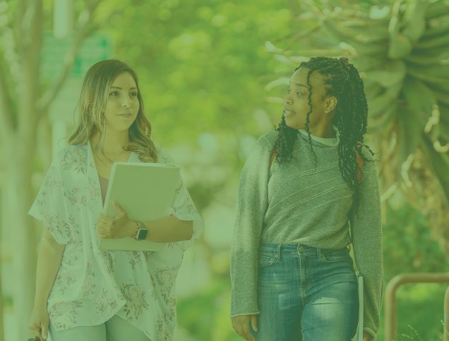 Two students holding books talk while walking down outdoor path