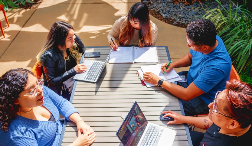 A group of students collaborate at a picnic table outdoors.