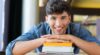 A man smiles at the camera while resting his hands on a stack of books and his chin on his hands.