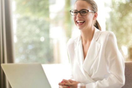 A smiling person wears glasses and a white blouse while seating at a desk with a laptop in front of them.