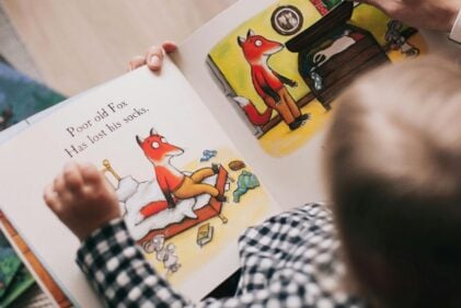 A young human child views a book with a picture of a cartoon fox upon it.